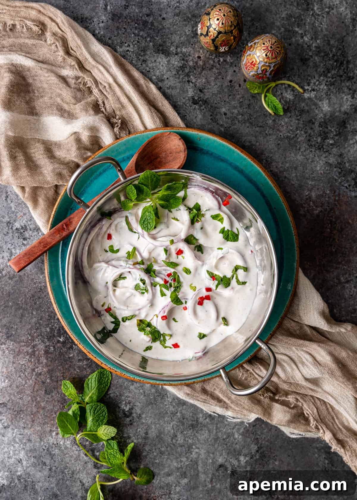 Overhead shot of a bowl of Onion Raita, garnished with fresh herbs and spices, on a rustic wooden table.