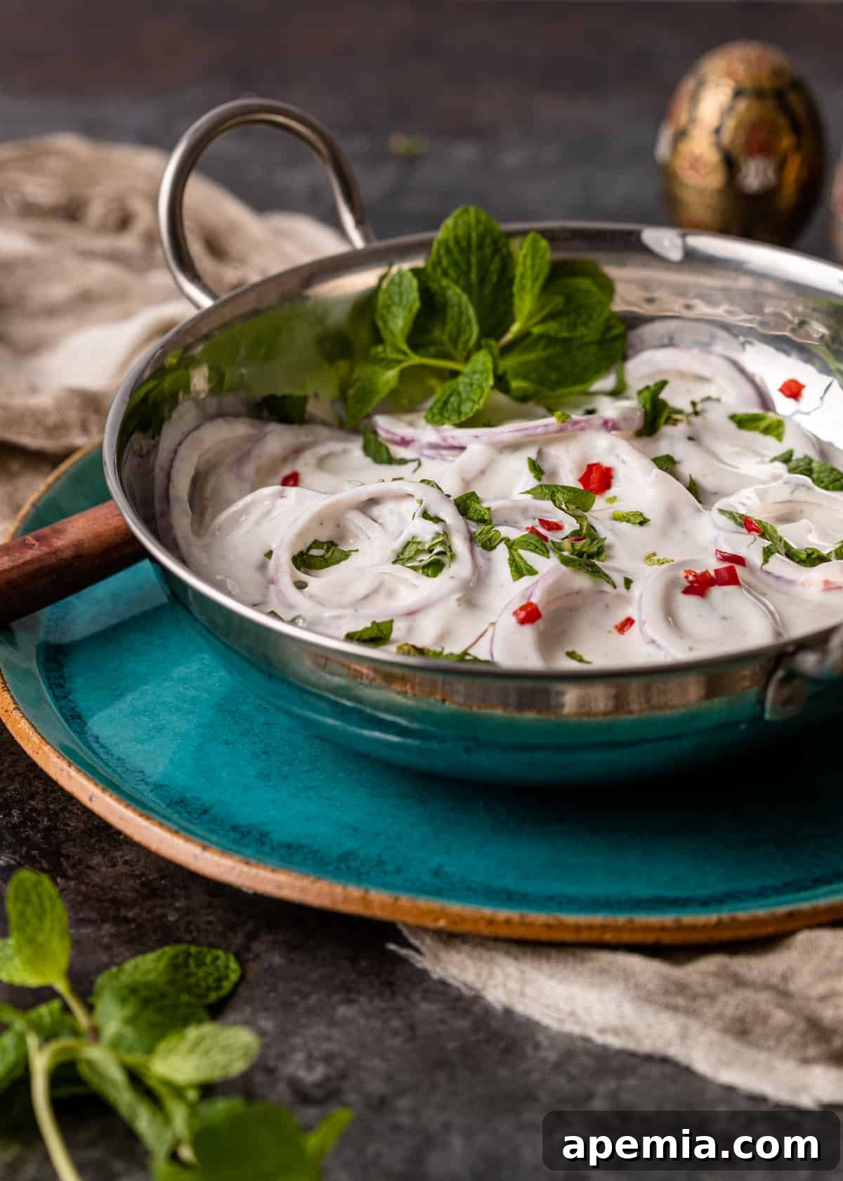 A beautifully presented bowl of Onion Raita, garnished with a sprig of fresh mint and a sprinkle of chili powder, on a light background.