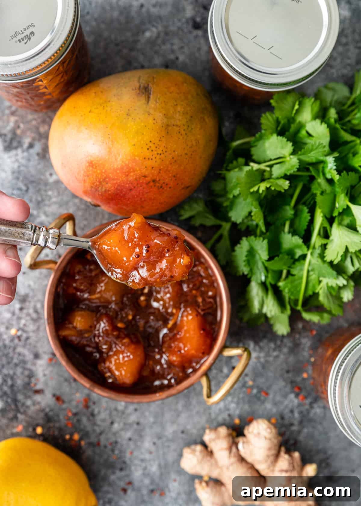 A spoon overflowing with golden mango chutney, held above a rustic copper bowl also brimming with chutney. A backdrop of fresh mangoes, spices, and other ingredients hints at the preparation process.