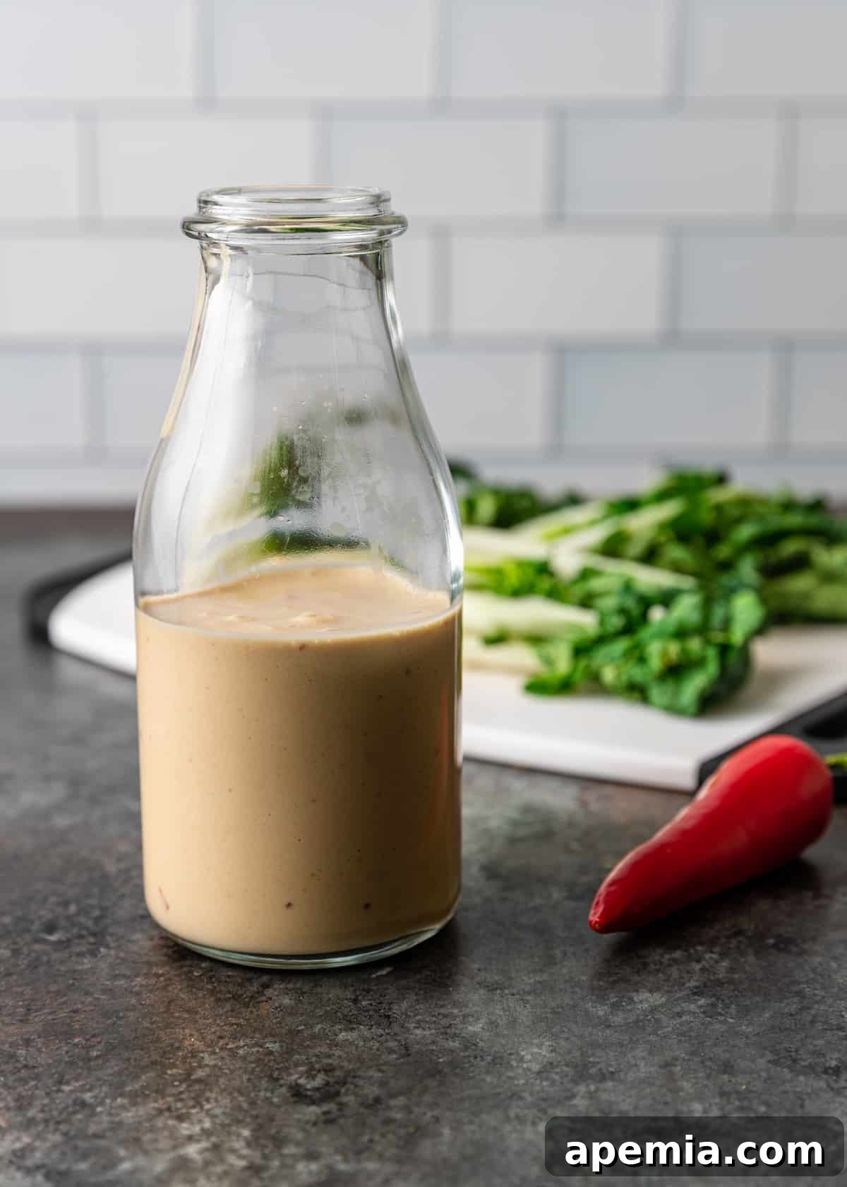 A large glass jar filled with freshly made Miso Tahini Dressing, with blurred greens and a red pepper in the background, suggesting its use in a salad.
