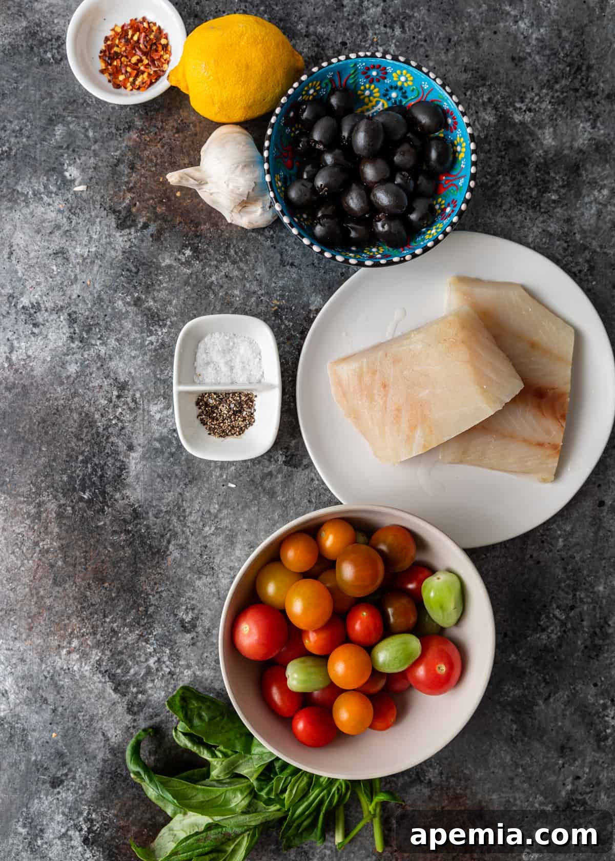 Overhead view of fresh ingredients laid out: cod fillets, cherry tomatoes, olives, basil, lemon, garlic, and spices.