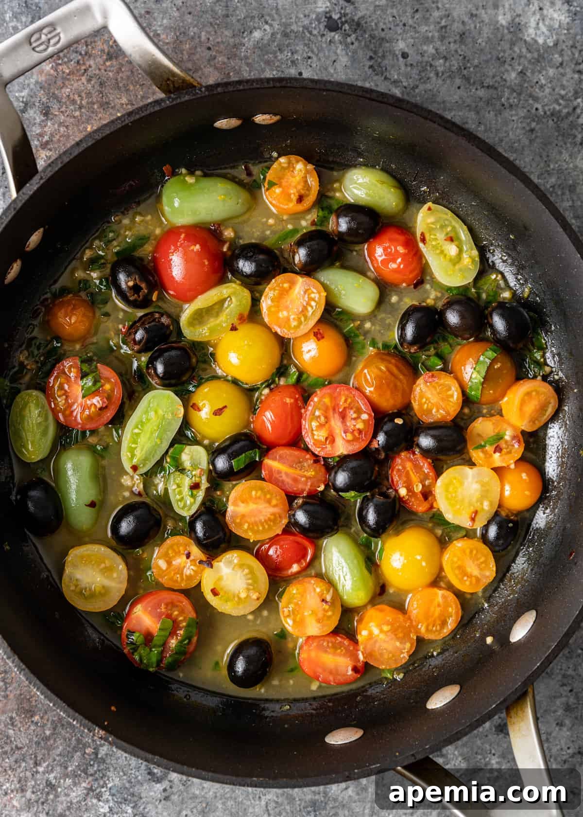 Overhead shot of cherry tomatoes, black olives, and herbs simmering in a skillet with a light sauce.