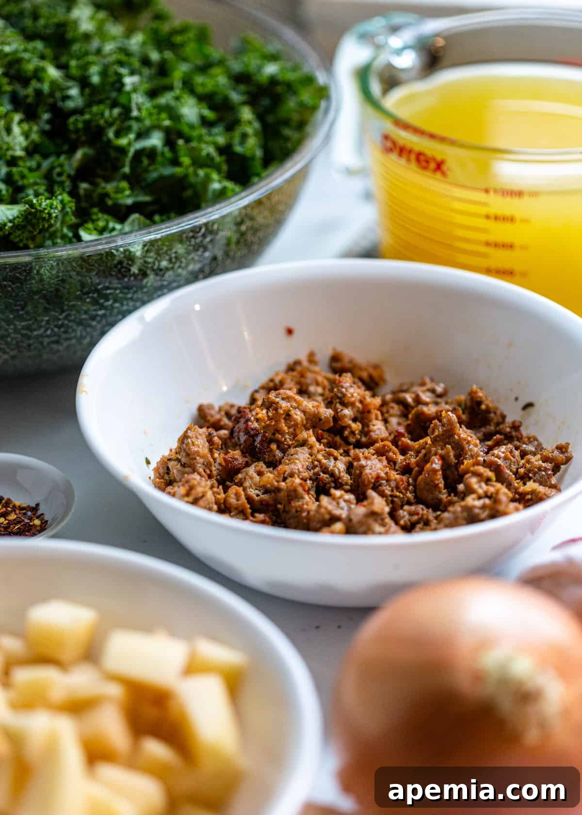 a white bowl full of cooked sausage with a bowl of kale and a pyrex measuring cup full of broth