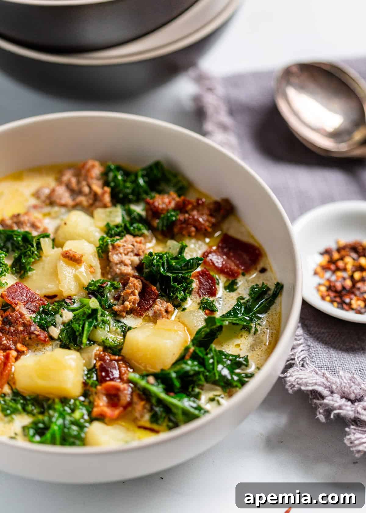 A white bowl full of zuppa toscana with a small bowl of red pepper flakes visible to the right