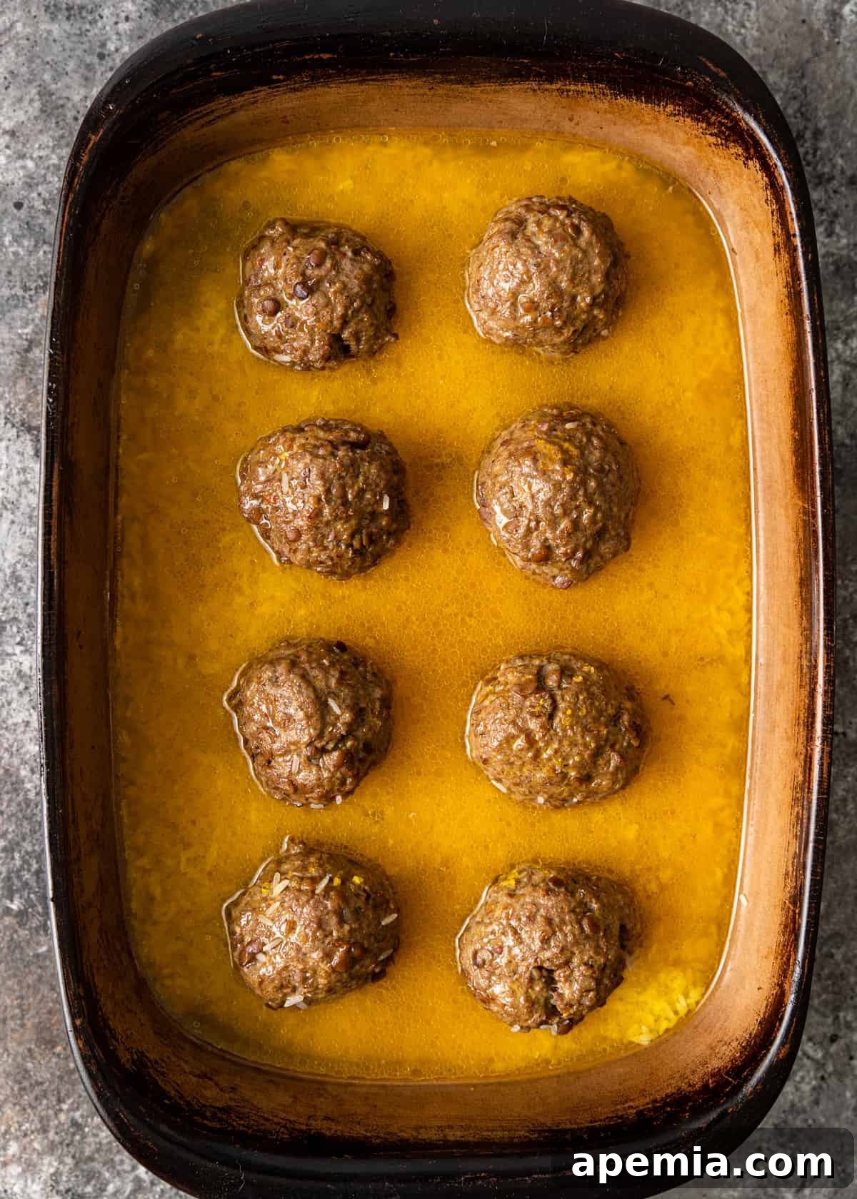 Mediterranean Lamb Lentil Meatballs and Zesty Lemon Rice 8 Overhead view of roasted lamb meatballs in a baking dish, with lemon-infused broth poured around them before adding rice.