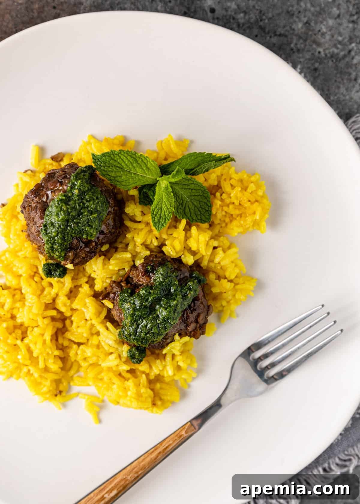 Mediterranean Lamb Lentil Meatballs and Zesty Lemon Rice 10 Overhead shot of two lamb meatballs artfully placed over lemon rice on a white plate, finished with a generous drizzle of mint pesto and a fork.