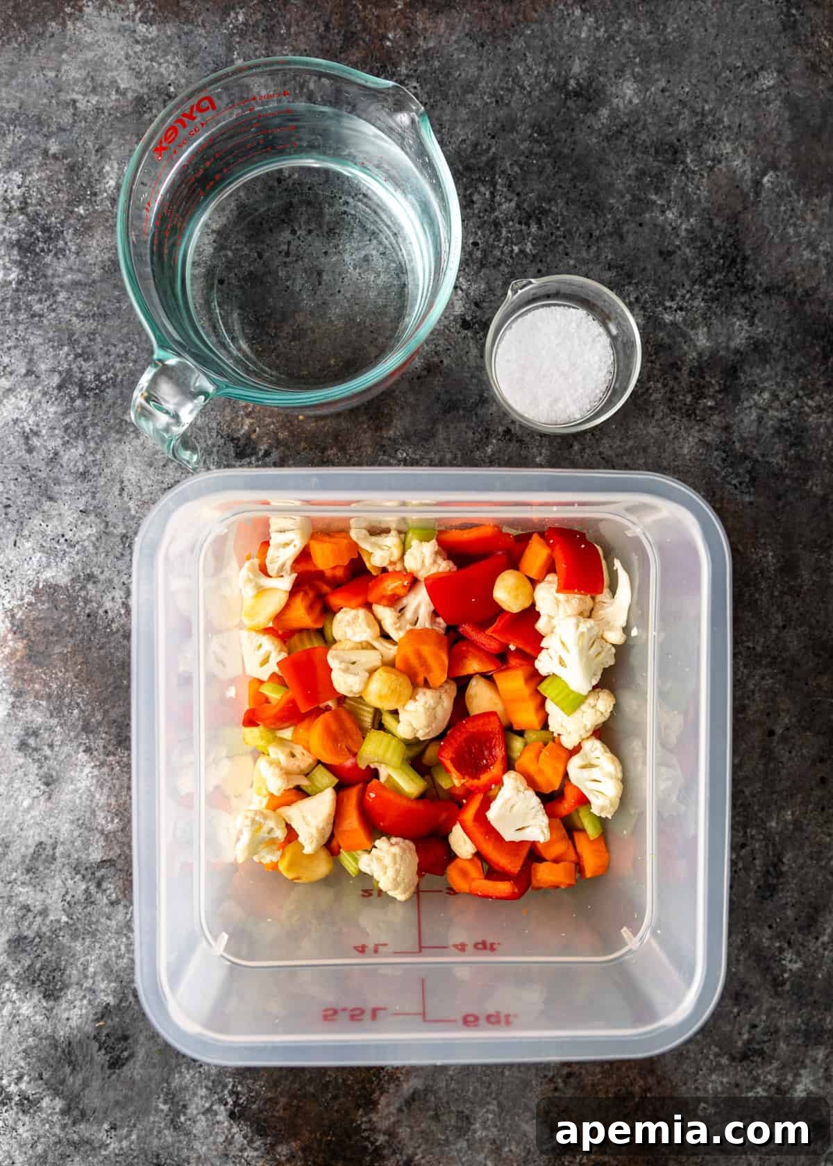 Overhead view of chopped vegetables neatly contained in a plastic bin, with a glass measuring cup of water and a small bowl of salt nearby, illustrating the initial preparation stages.