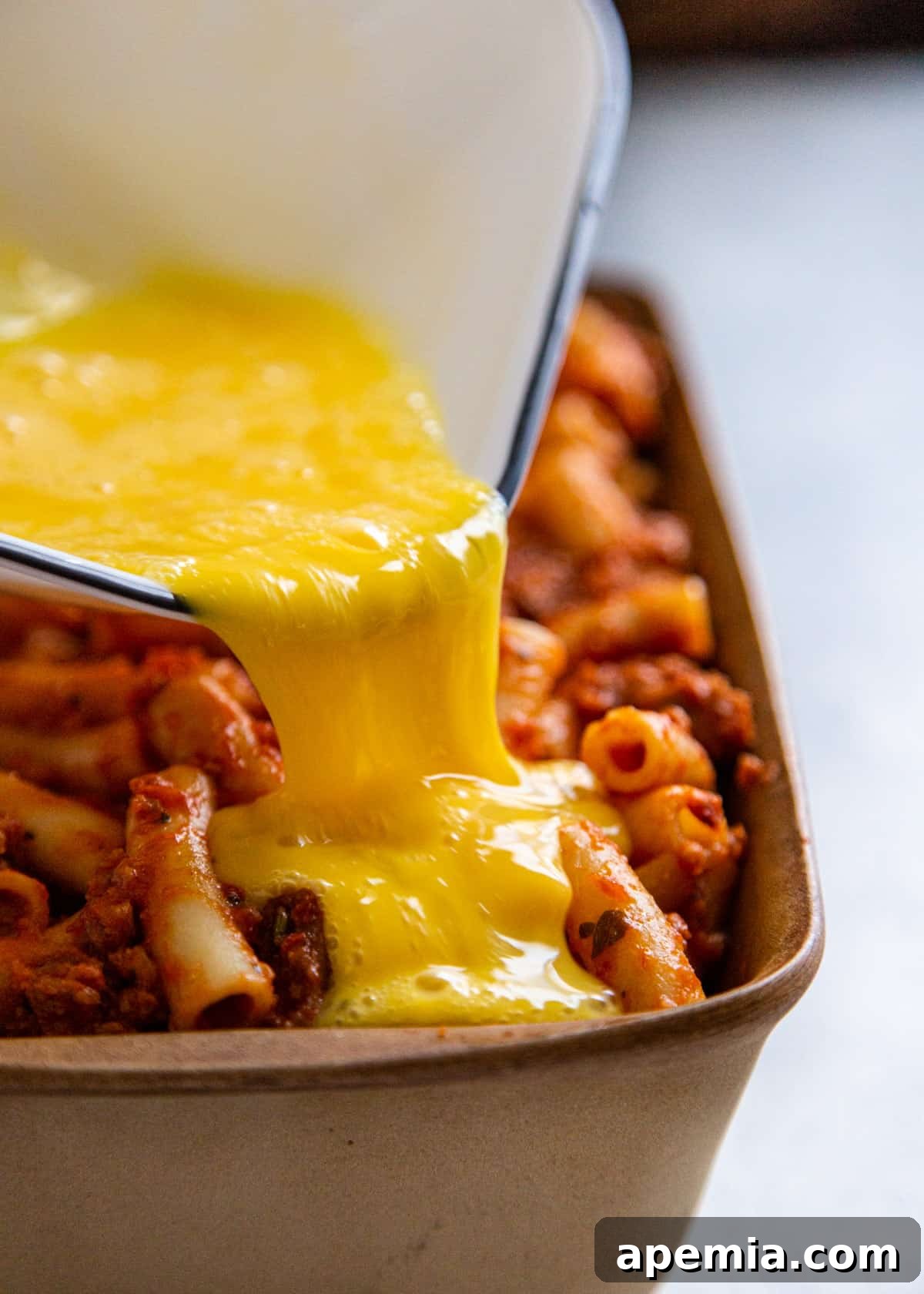 Side view of someone pouring melted cheese sauce over baked ziti in a pan