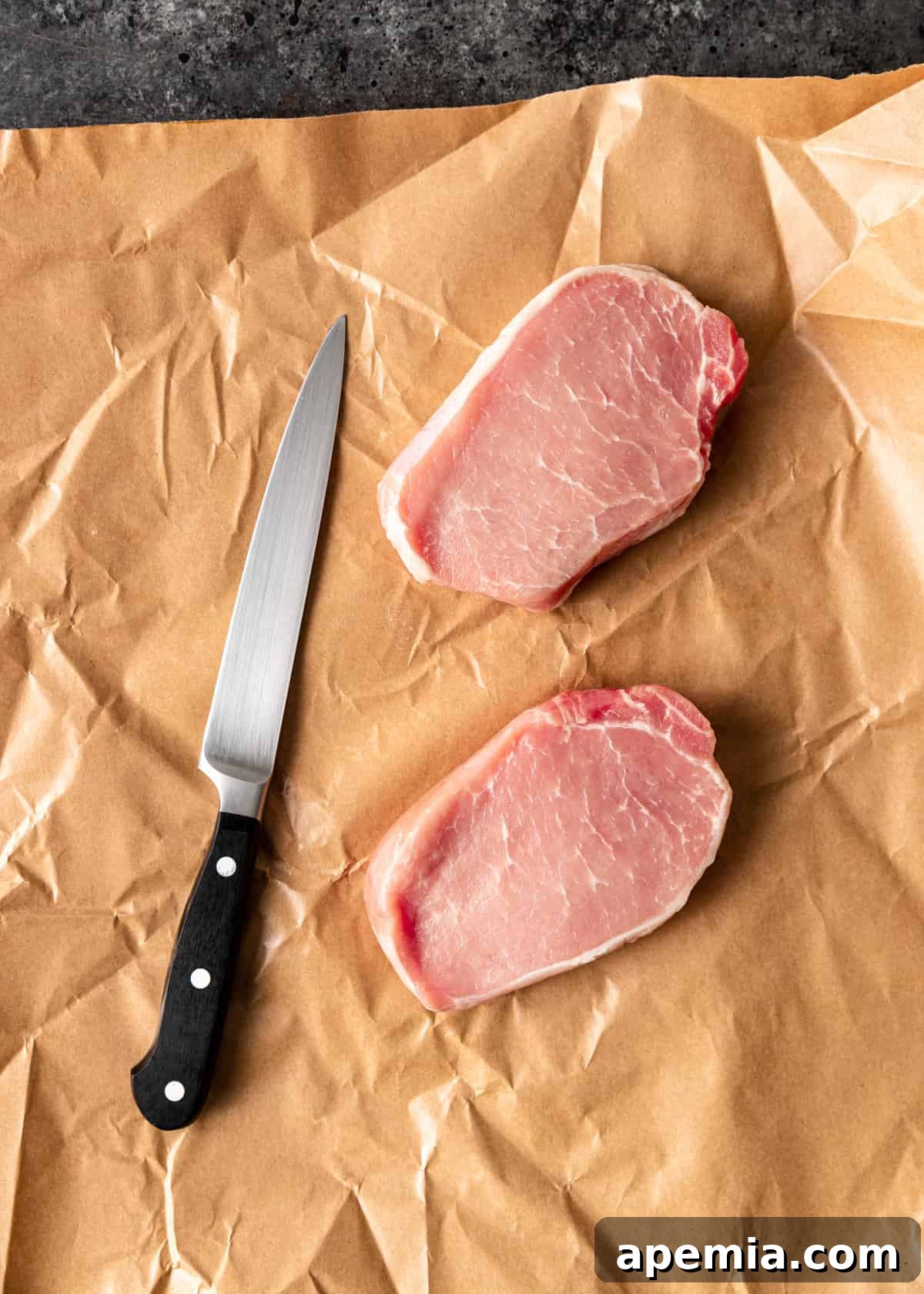 Overhead view of two thick pork chops on butcher paper, with a knife positioned to show how to butterfly them for tonkatsu.
