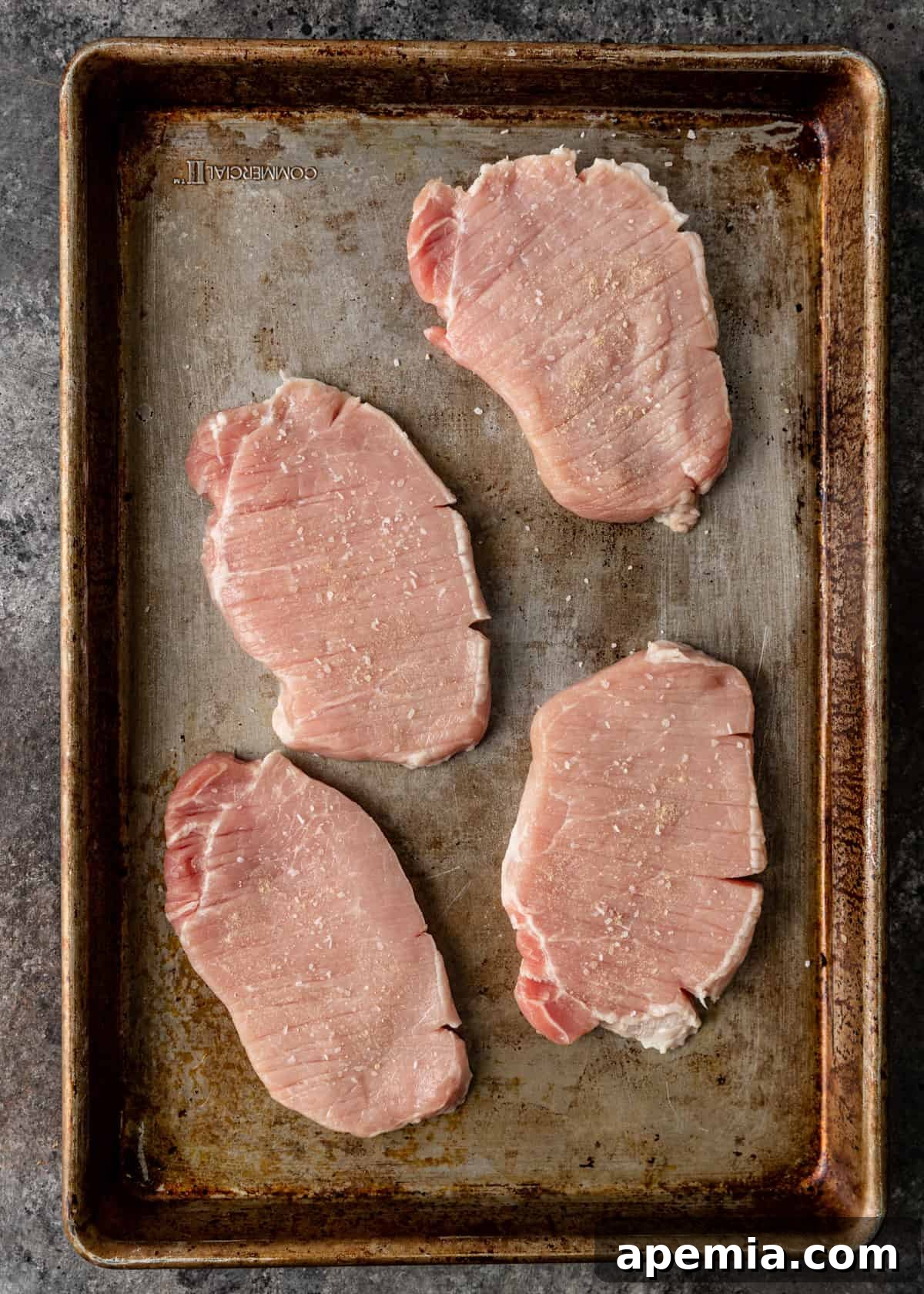 Overhead shot of four seasoned and tenderized pork cutlets laid out on a baking sheet, ready for the next step of breading.