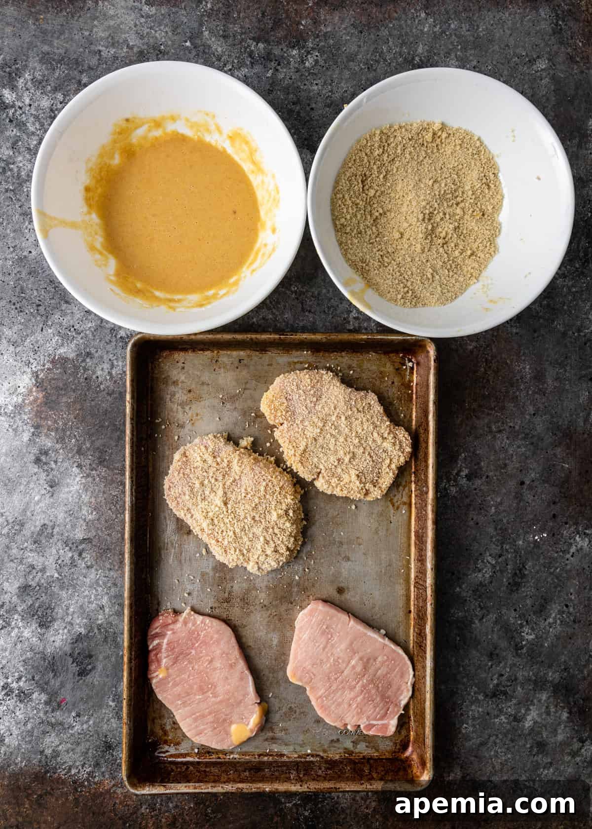 Overhead view of a dredging station: a baking sheet with four pork cutlets, two of which are already perfectly breaded with panko, ready for frying.