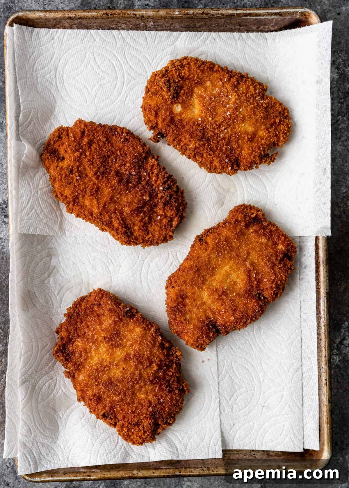 Overhead view of several freshly cooked pork katsu cutlets resting on paper towels on a baking sheet, allowing excess oil to drain.