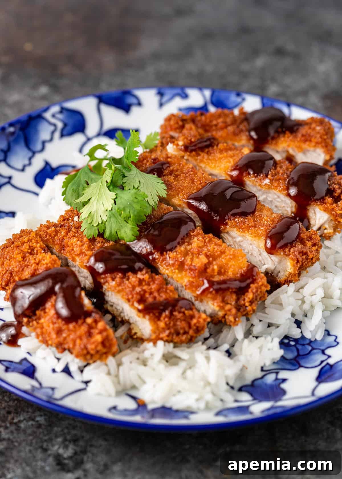 Closeup of freshly sliced pork katsu, drenched in katsu sauce and garnished with parsley, elegantly served over white rice on a blue and white patterned plate.