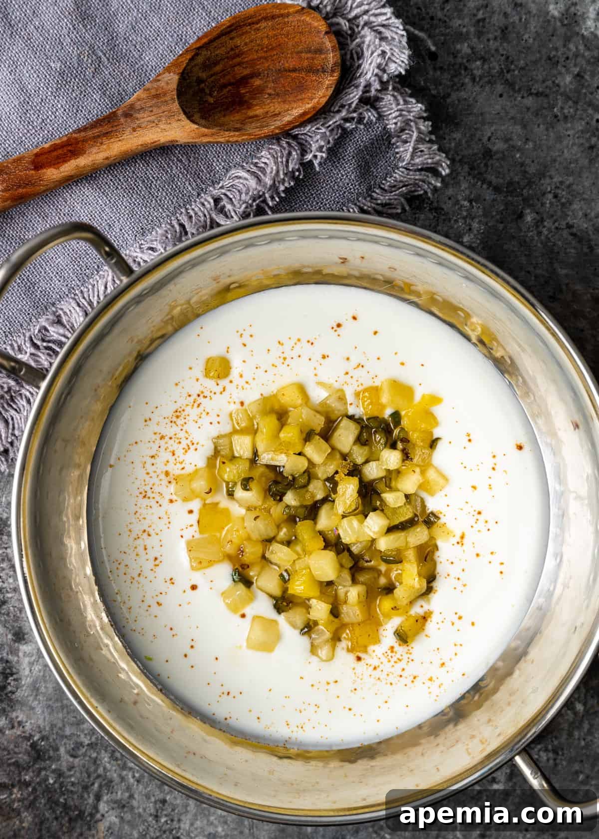 overhead: pineapple pachadi in a metal pan on a gray countertop with a blue towel and wooden spoon visible