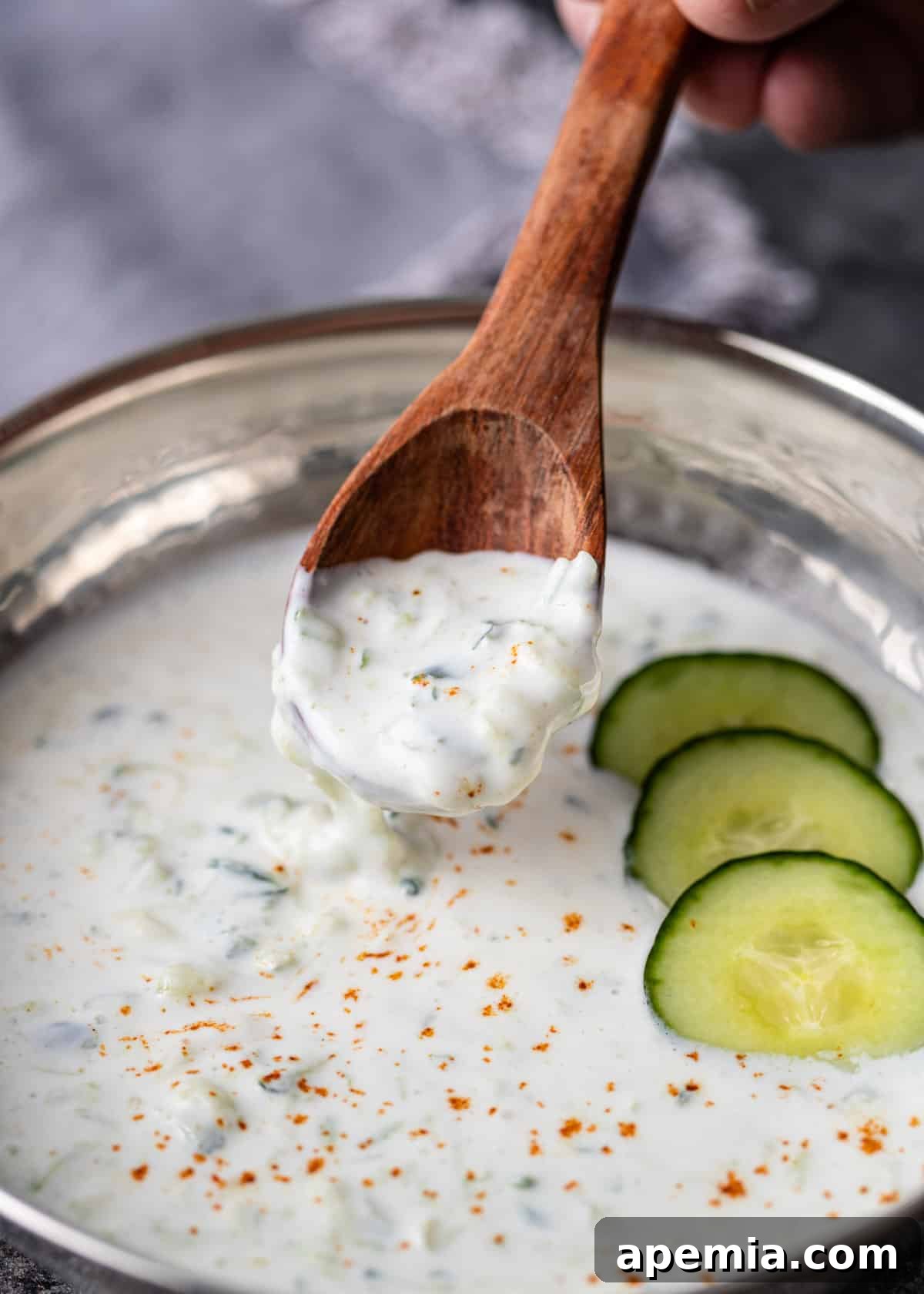 closeup: a bowl of cucumber raita with sliced cucumbers on top with a wooden spoon scooping out the yogurt sauce