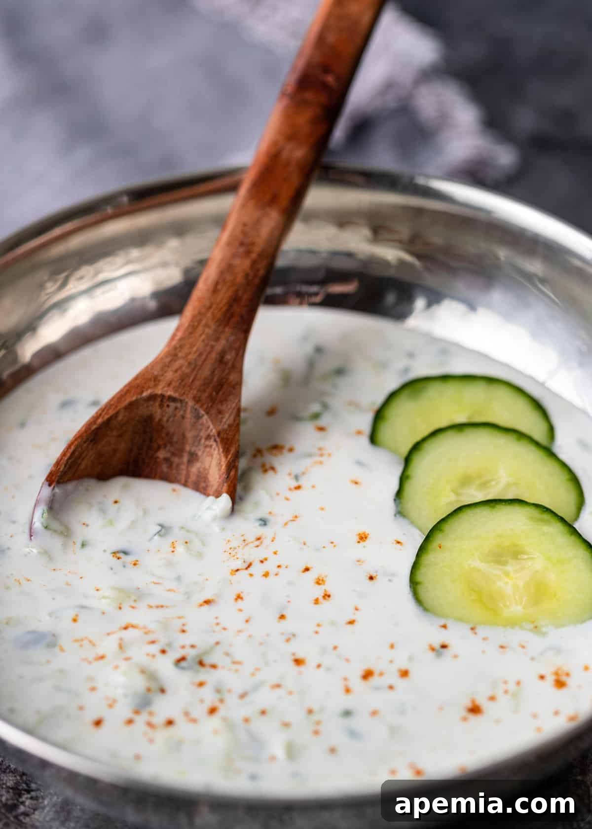 closeup: a wooden spoon in a bowl of cucumber raita with sliced cucumbers on top, showcasing the dish's texture
