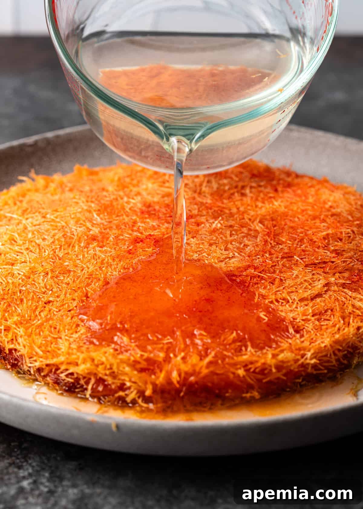 Close-up shot of sweet sugar syrup being poured generously over the hot, freshly baked knafeh pastry.