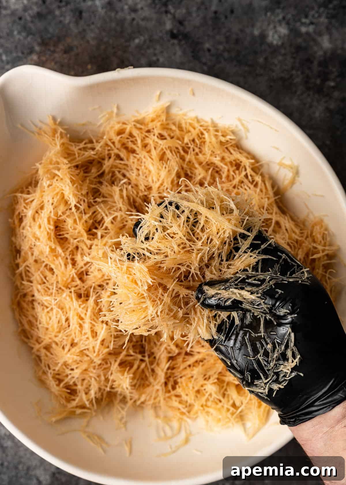 Overhead view of a black gloved hand meticulously mixing shredded filo dough (kataifi) with golden melted ghee in a large bowl.