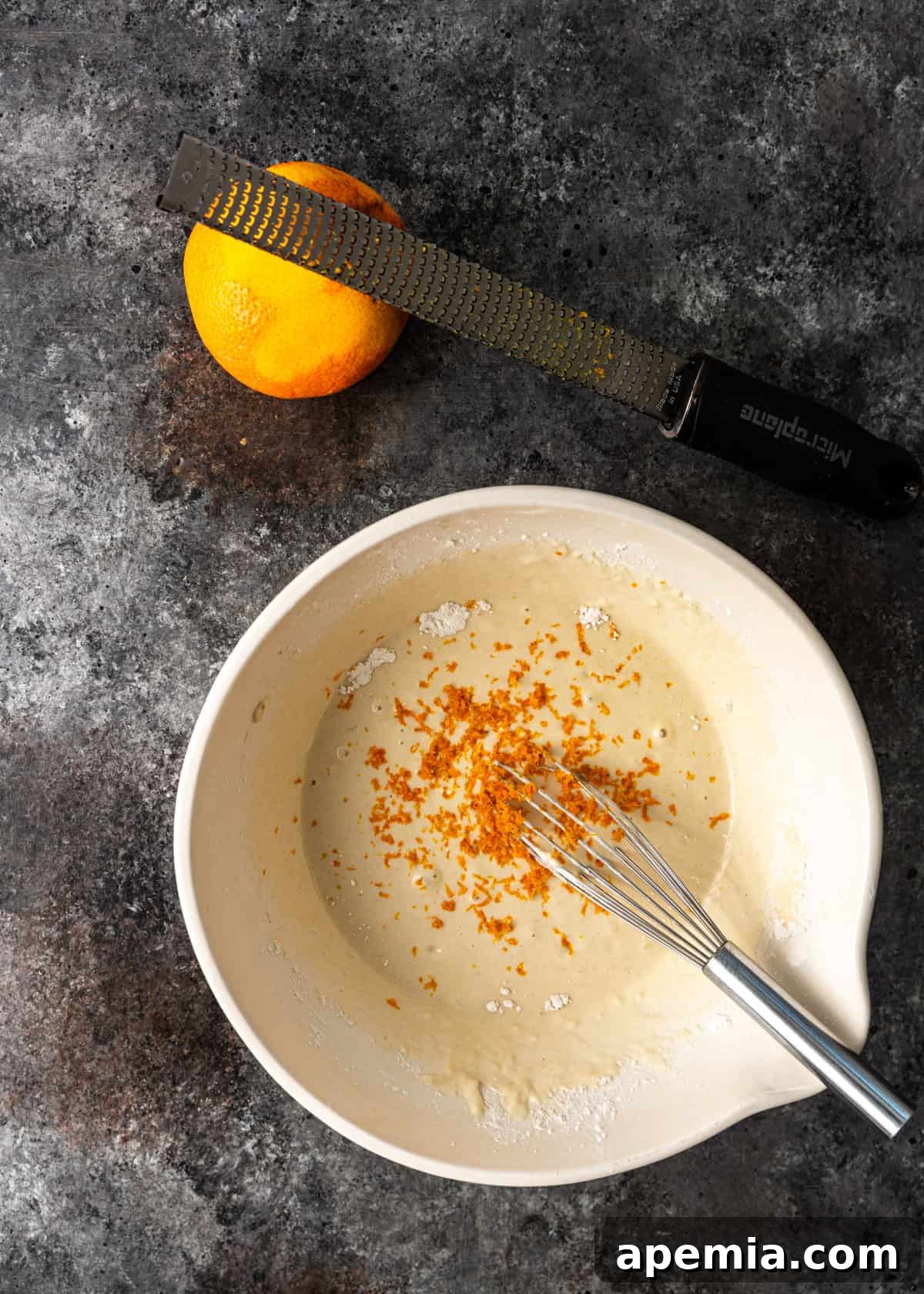 Overhead shot of a bowl with orange chicken batter being smoothly mixed with a whisk, showing the incorporation of orange zest.