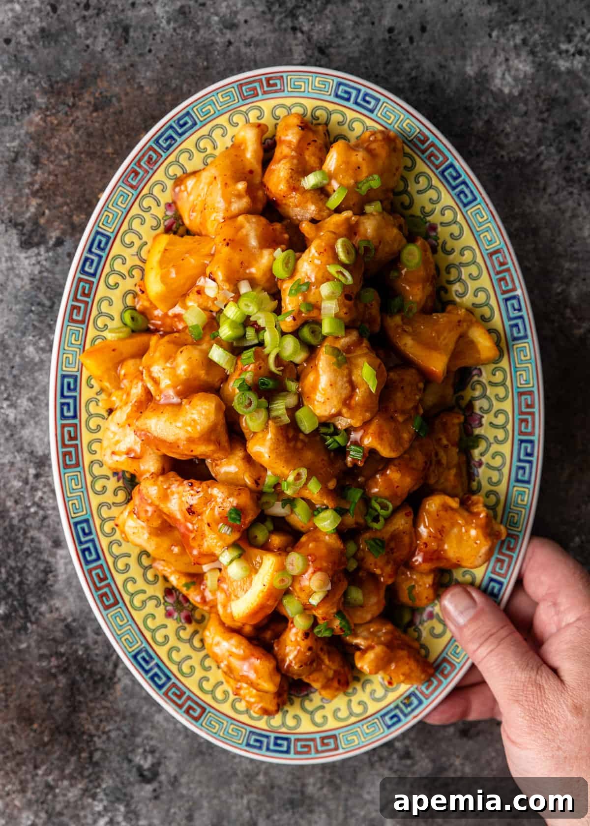 Overhead shot of a man's hand holding a large platter filled with freshly prepared orange chicken, garnished with sliced green onions and orange segments.