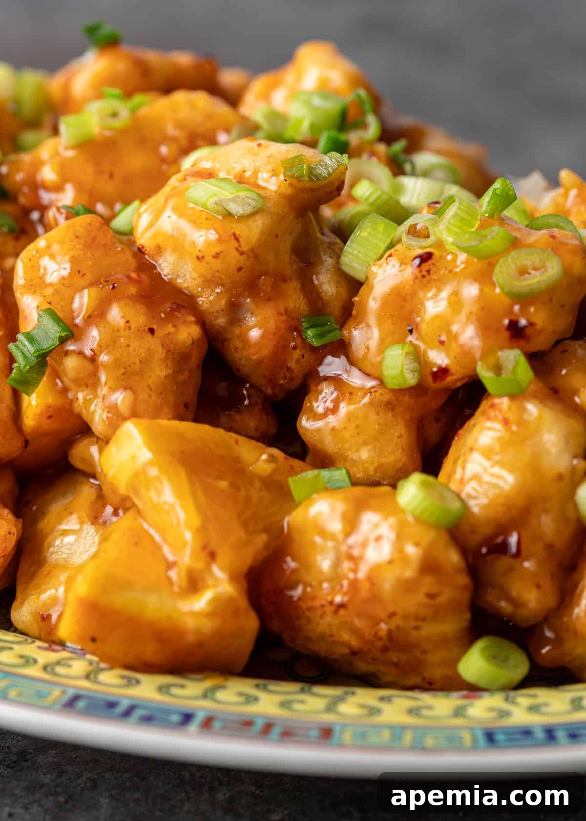 Extreme closeup of an individual piece of orange chicken on a ceramic plate, showing the texture of the crispy coating, sticky sauce, and a fresh green onion garnish.