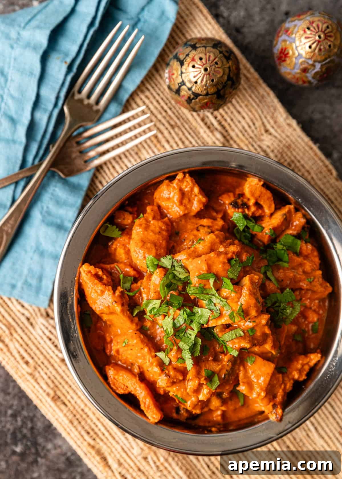overhead: a metal bowl full of chicken tikka masala with forks and a blue napkin to the side