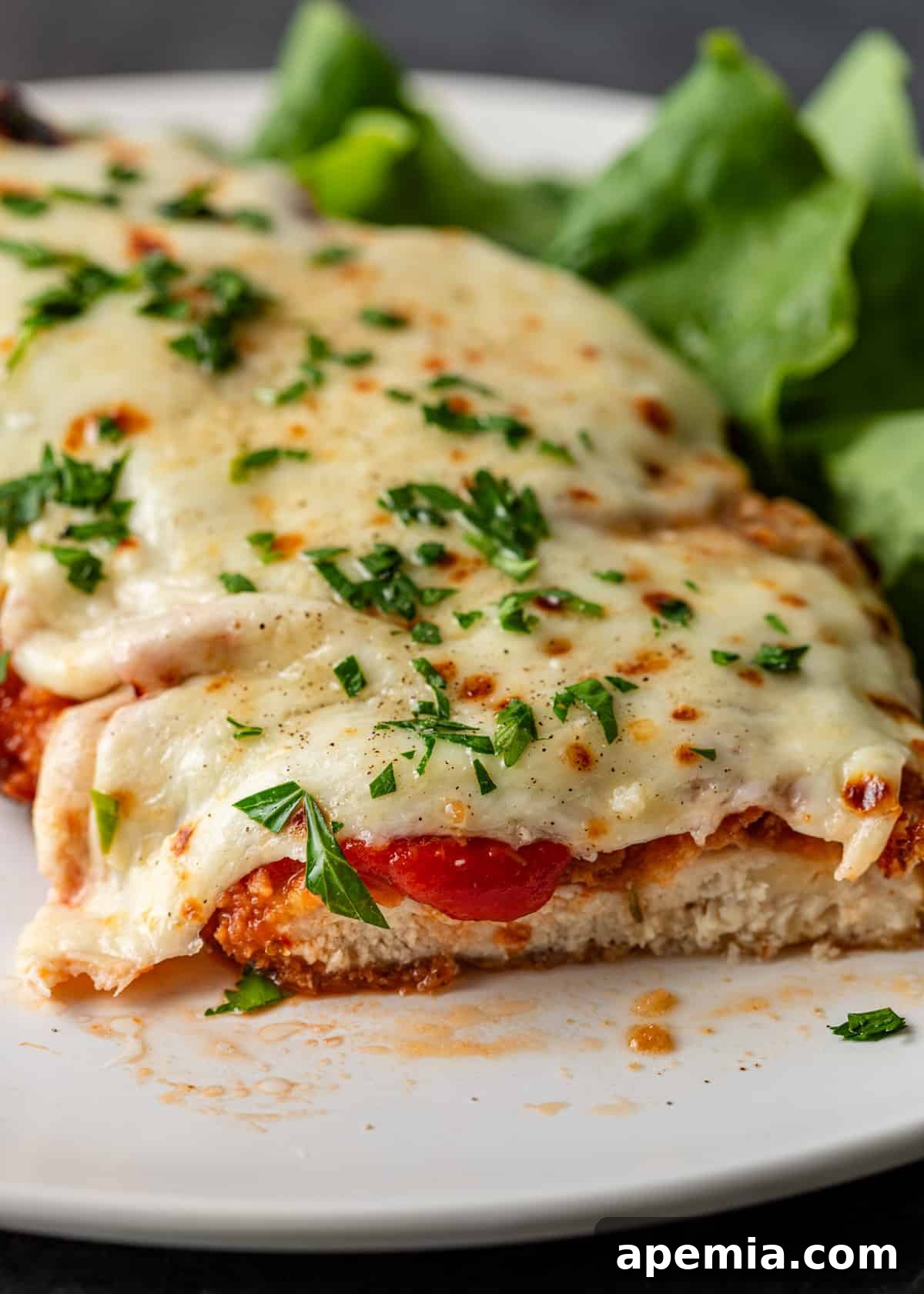 closeup: chicken parmigiana cut on a plate so that the chicken is visible. It's covered with marinara, cheese, and fresh herbs