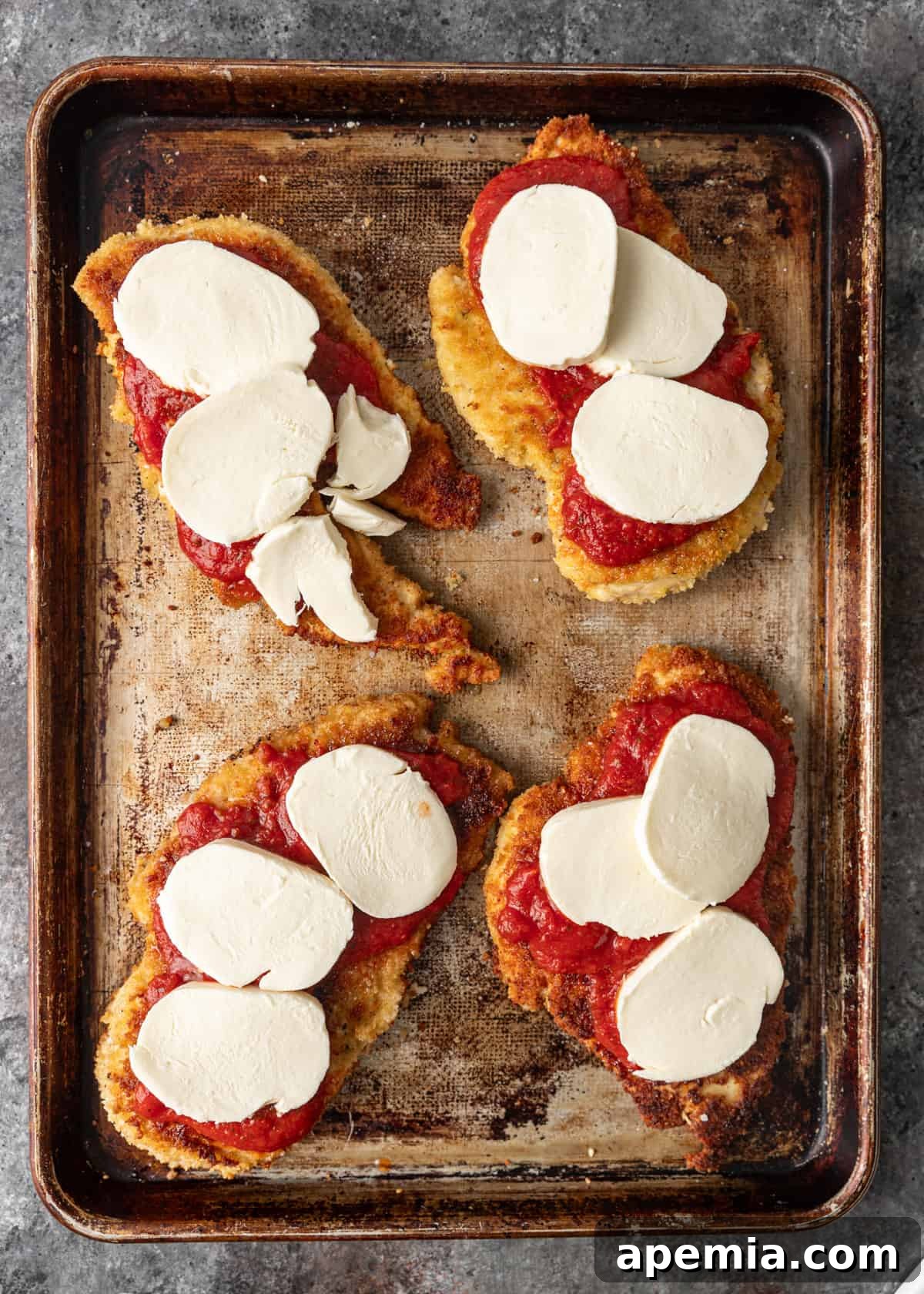 overhead: cooked breaded chicken breasts on a baking sheet with marinara and mozzarella on top