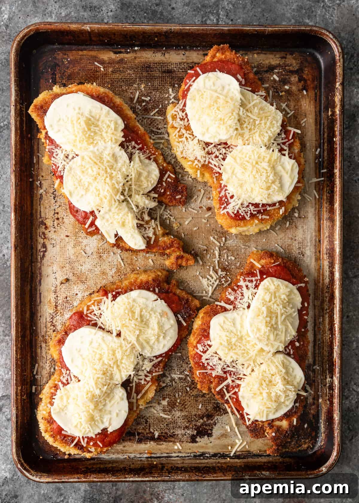 overhead: cooked breaded chicken breasts on a baking sheet with marinara, mozzarella, and parmesan on top