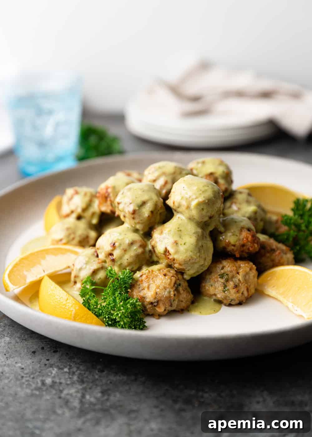A beautifully presented stack of chicken meatballs, garnished with creamy Alfredo sauce, fresh lemon, and parsley on a plate.