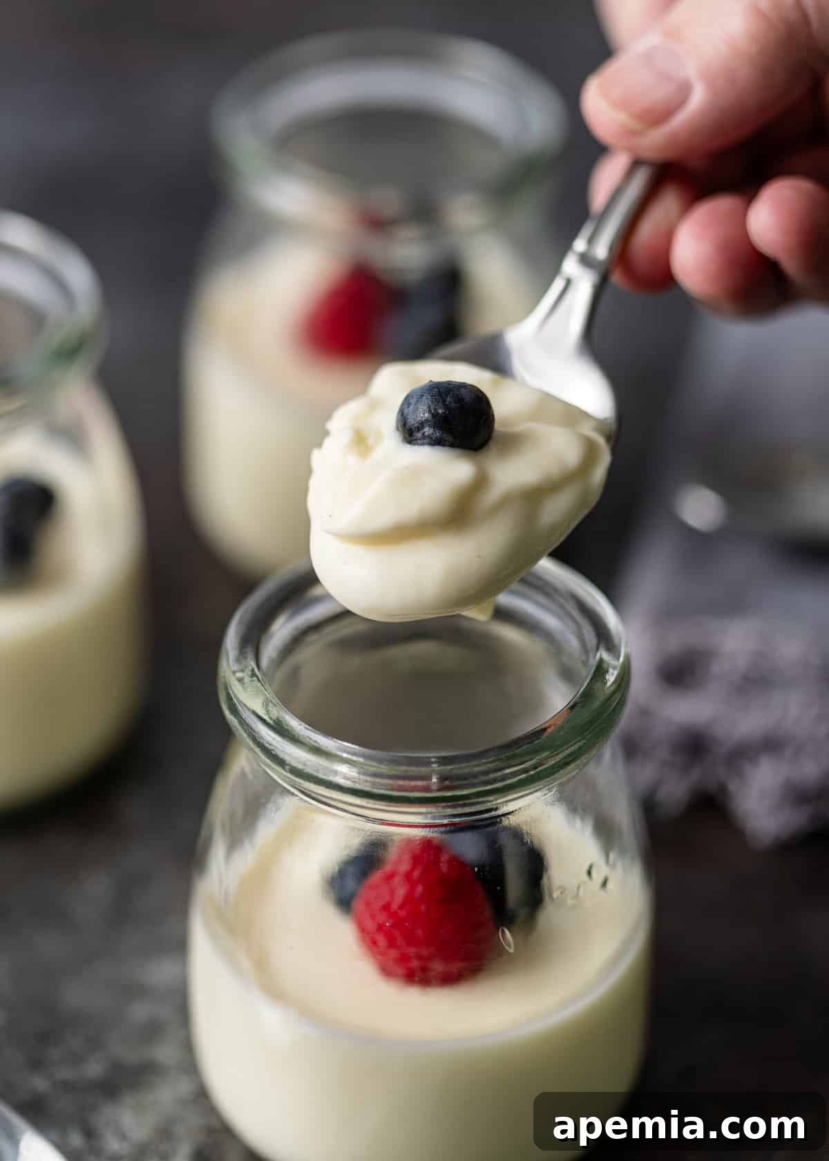 Zesty Creamy Lemon Posset 9 Closeup of a hand lifting a spoonful of lemon posset, topped with a single fresh blueberry, from a glass container.