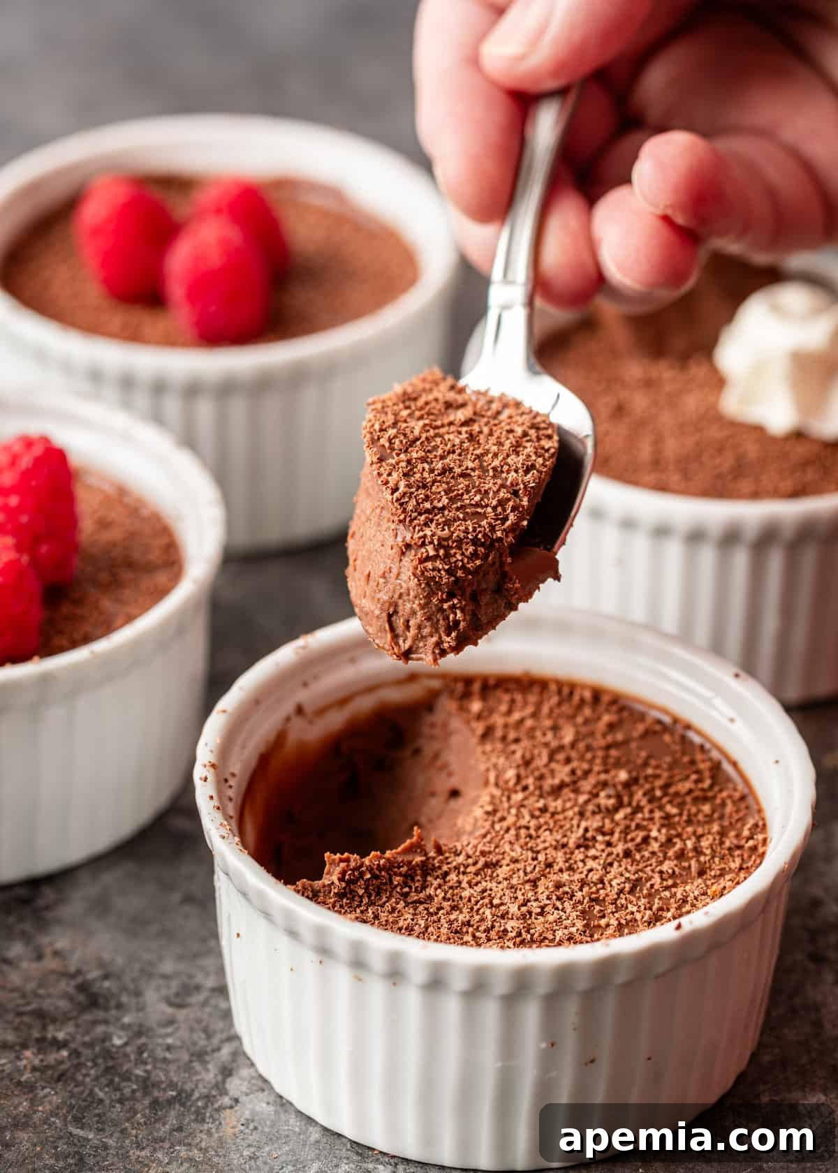 Decadent Chocolate Pots de Crème 8 closeup: a hand holding a spoonful of dark chocolate pot de creme, hovering over a white ramekin with more delicious dessert inside, showing its creamy texture