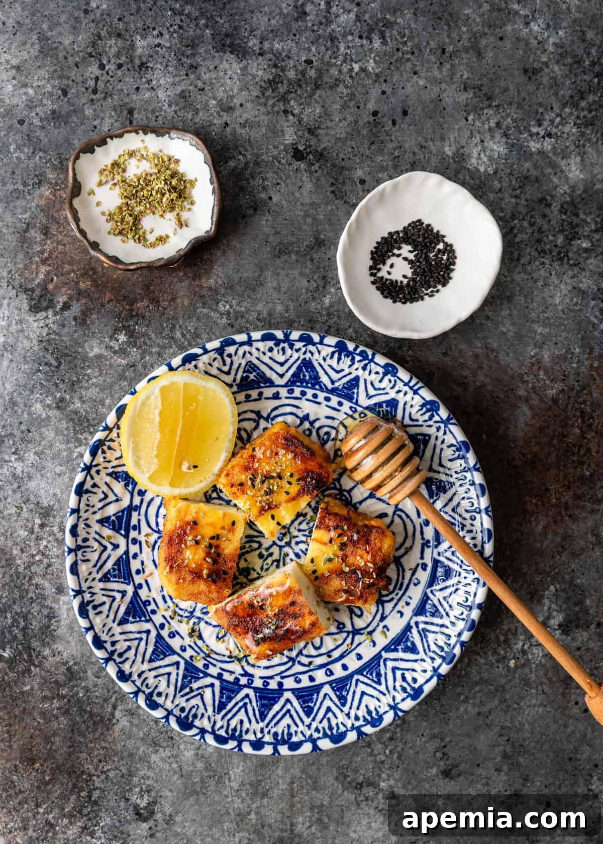 Fiery Greek Cheese Saganaki 3 Overhead view of Saganaki served on a blue and white ceramic plate, adorned with honey and a lemon wedge, with two small bowls of fresh herbs positioned to the side.