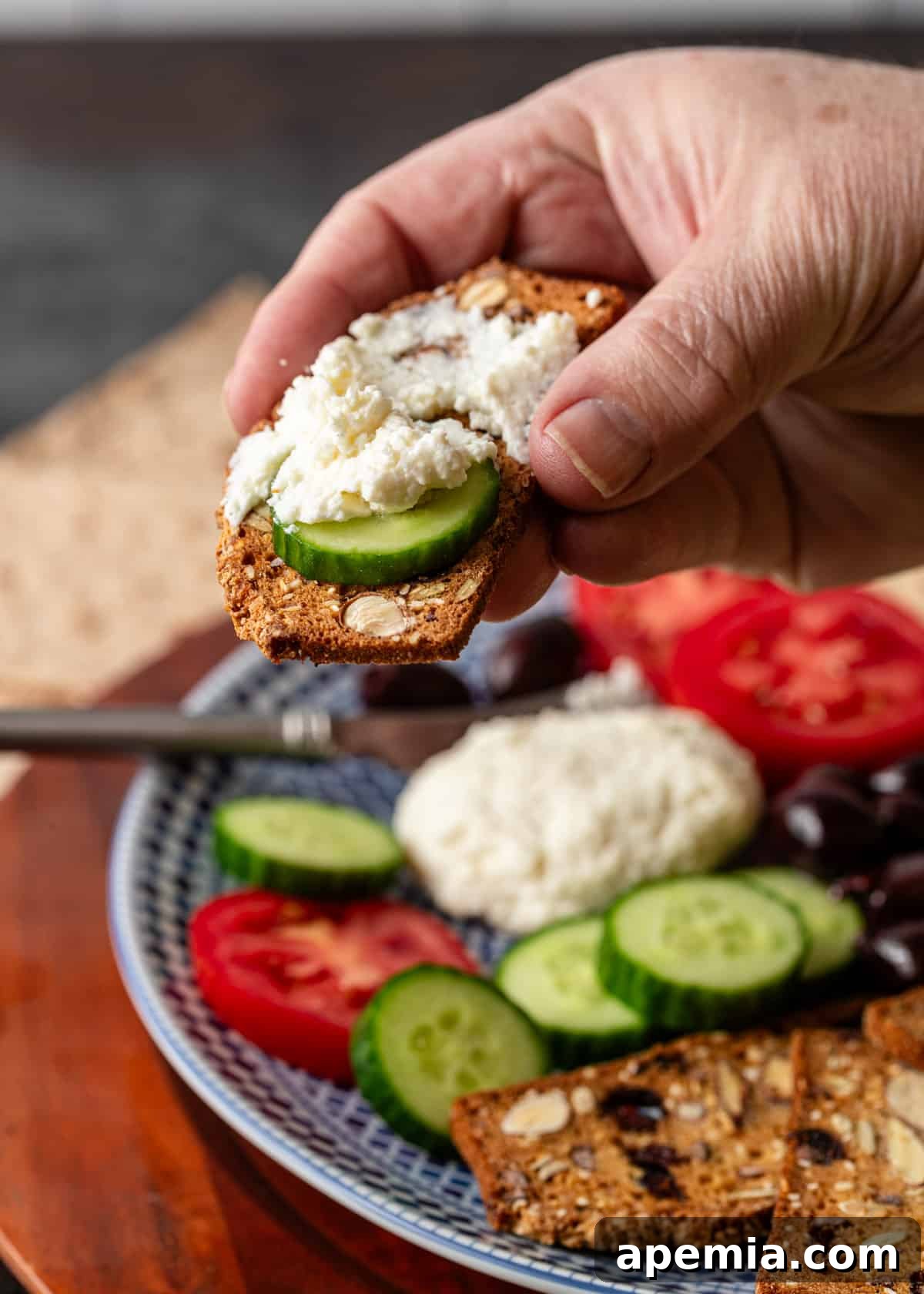 A hand lifting up a cracker topped with a fresh cucumber slice and a delectable piece of Joban cheese.