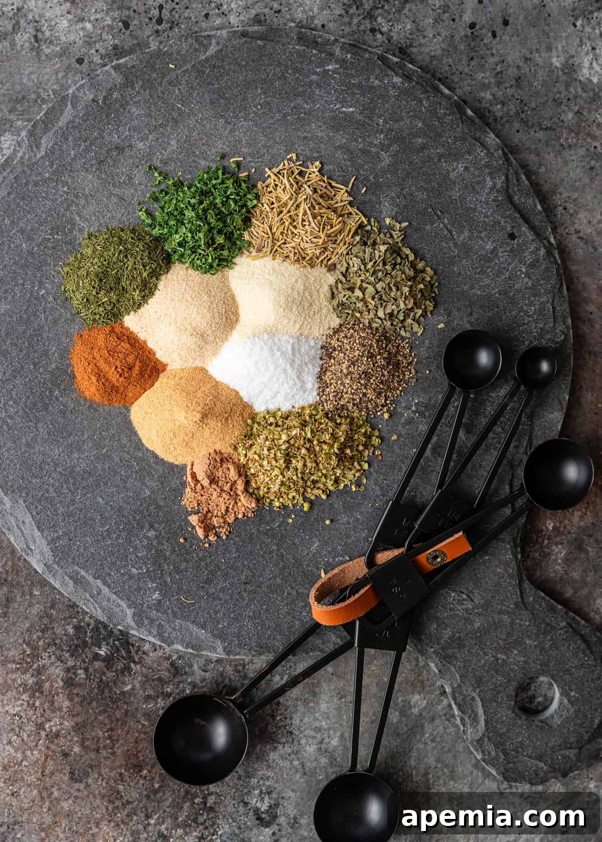 An overhead shot showcasing an array of individual spices and herbs, artfully arranged on a slate board alongside measuring spoons, illustrating the components of a Greek seasoning blend.