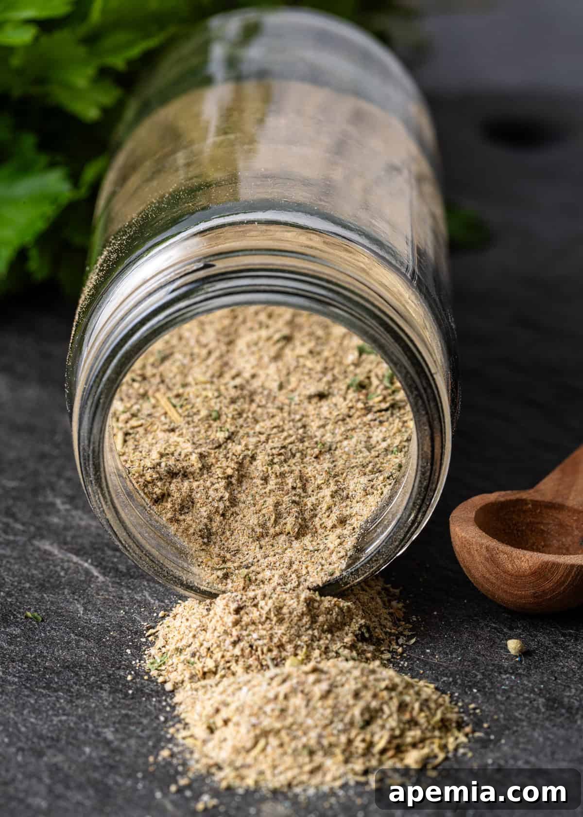 A close-up of a small glass jar filled with Greek seasoning, with some of the blend elegantly spilling onto a dark slate counter, illustrating its inviting texture.