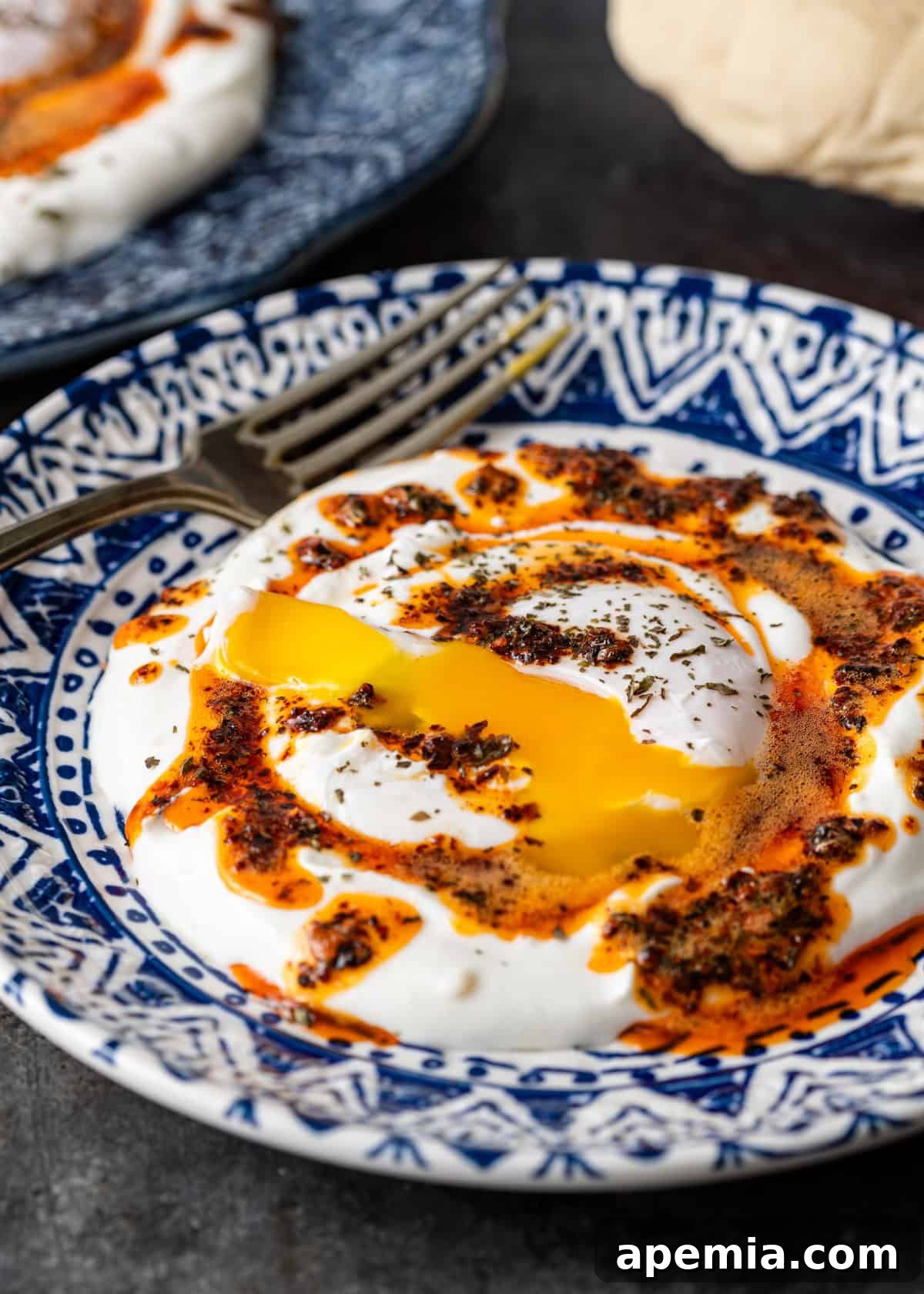 Close-up of a Cilbir dish on a blue ceramic plate, highlighting the perfectly runny yolk and the rich chili butter cascading over the poached egg and creamy yogurt.