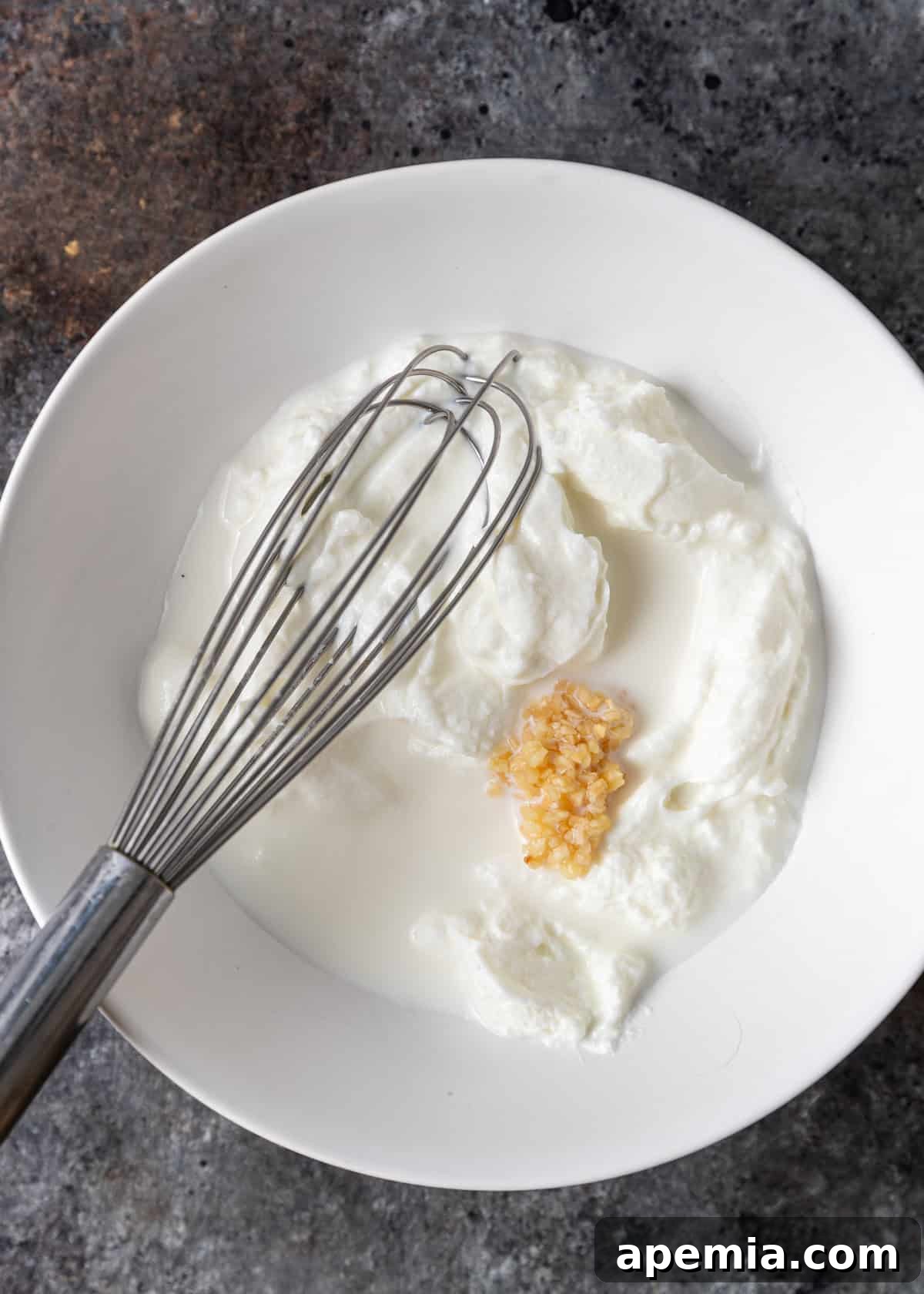 Overhead process shot demonstrating how to prepare the creamy garlic yogurt sauce for Çilbir, with ingredients laid out.