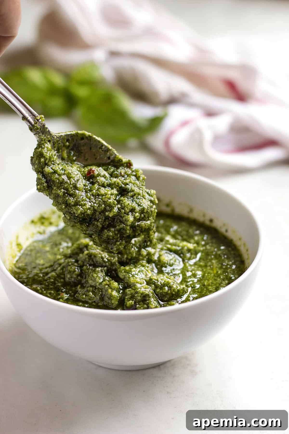 Close-up of a spoon lifting fresh basil pesto from a white bowl, showing its vibrant green color and chunky texture