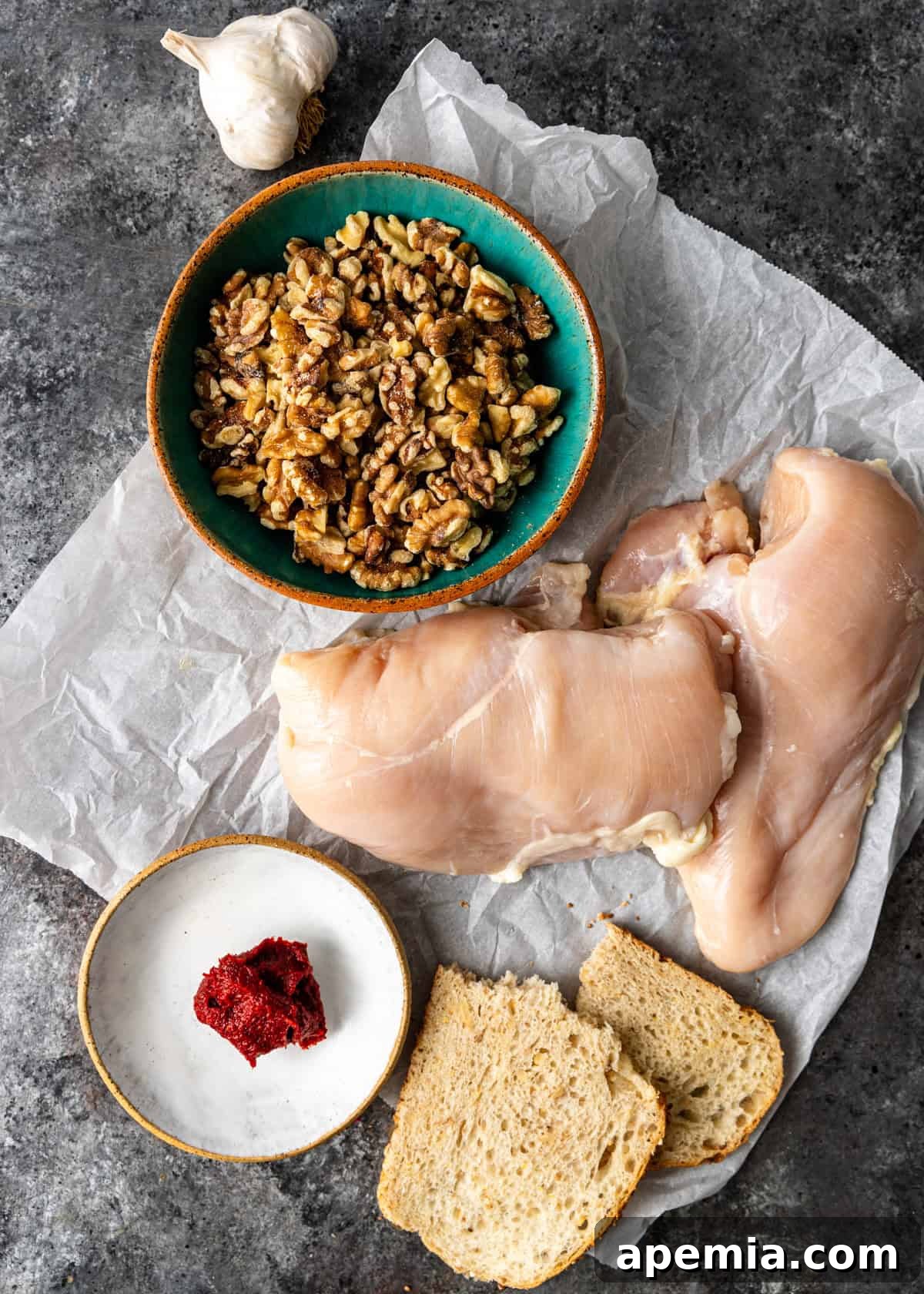 Overhead view of all fresh ingredients laid out for preparing Turkish chicken salad, including chicken breasts, walnuts, garlic, bread, cilantro, and red pepper paste.