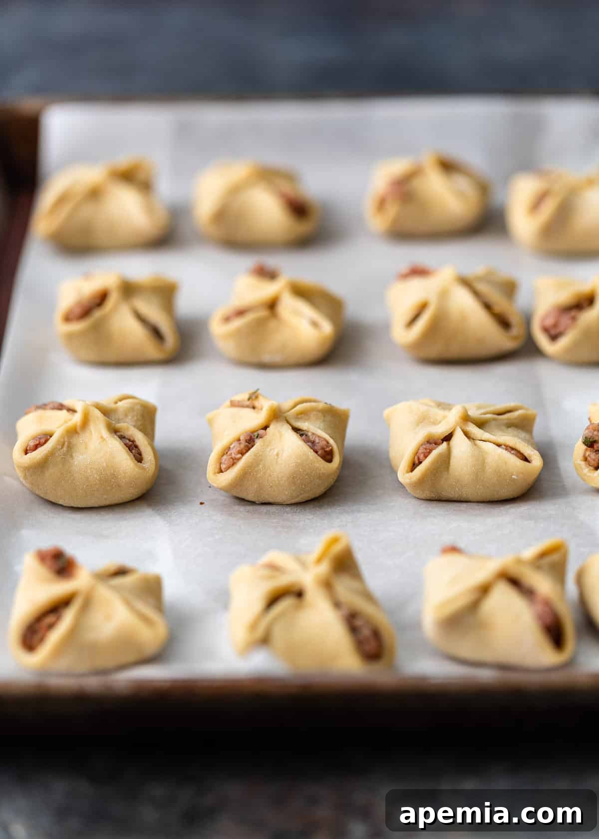 Side view of several uncooked Manti dumplings neatly arranged on a baking sheet lined with parchment paper, awaiting their turn in the pot.