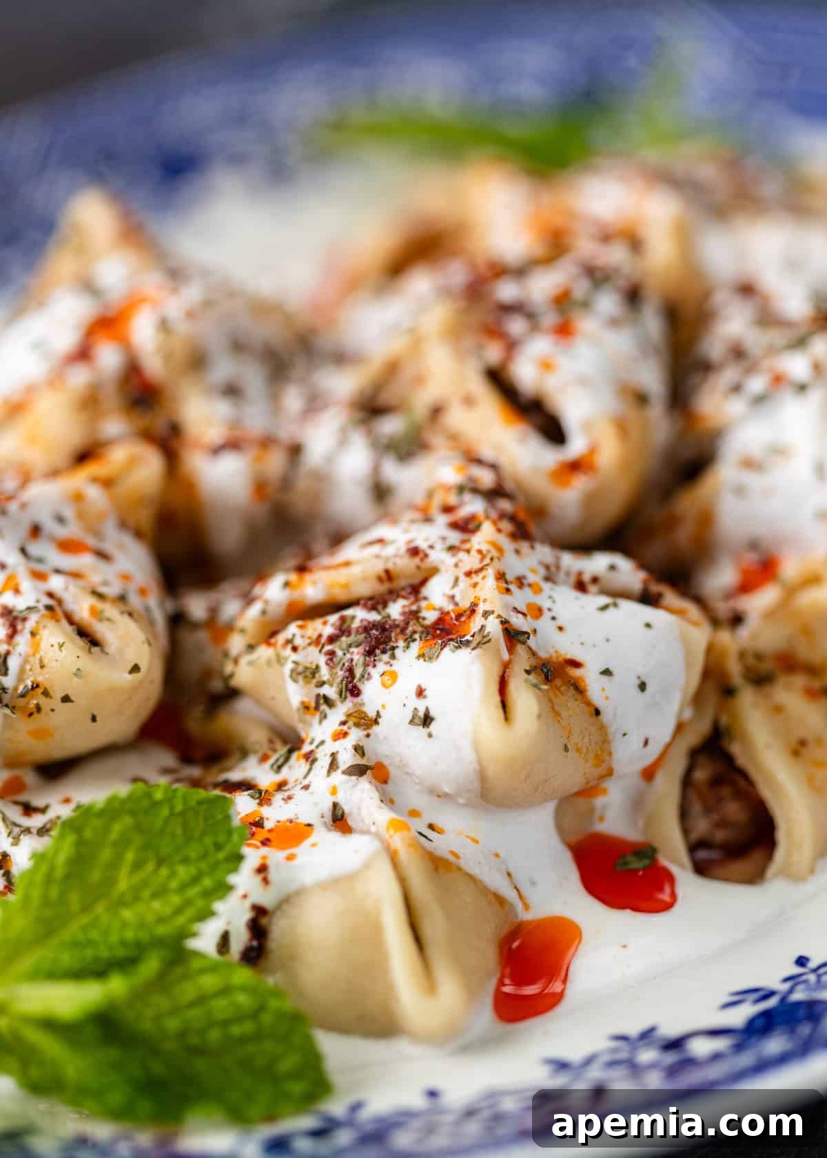 Close-up shot of a platter of Turkish dumplings generously covered in creamy white sauce, with a vibrant chili drizzle and fresh herbs for garnish.