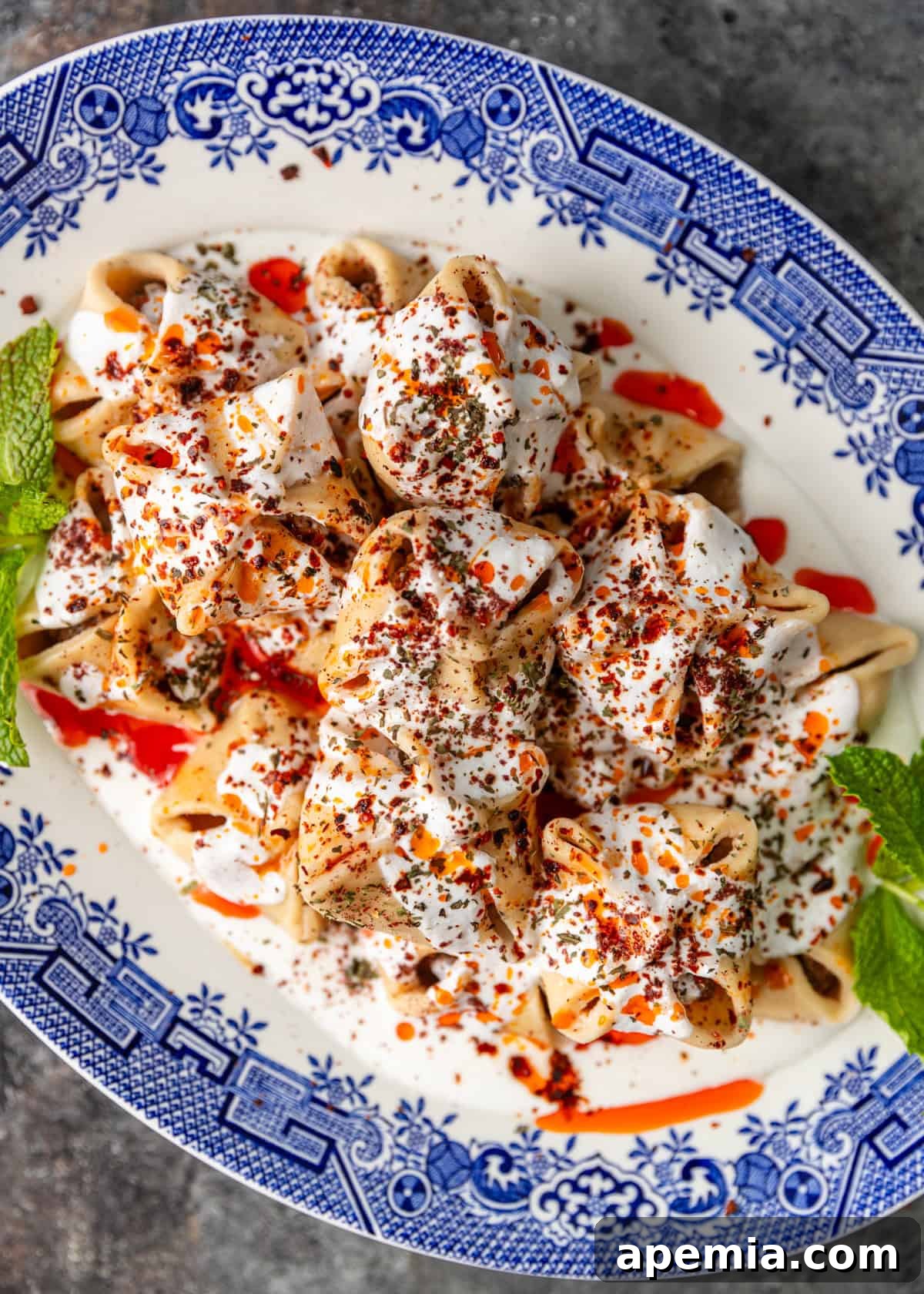 Overhead view of a large platter of homemade Turkish Manti, generously topped with creamy white yogurt sauce and drizzled with spicy red chili oil.