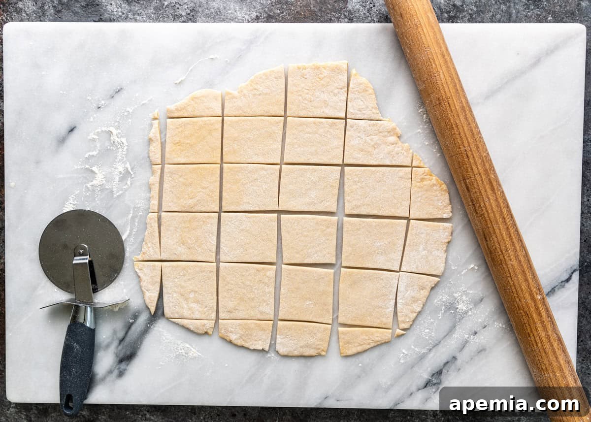 Overhead process shot: Hands using a pizza cutter to precisely cut the rolled-out dough into small squares for making Manti dumplings.