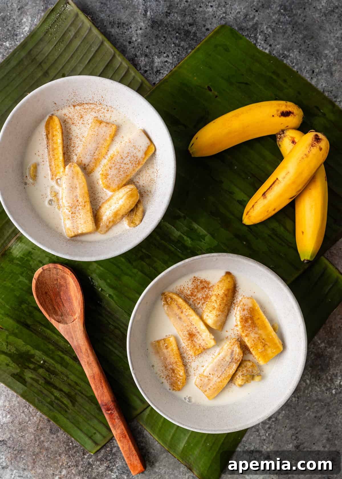 Overhead shot of two white bowls of Chak Ktiss (banana in coconut milk) garnished with cinnamon, placed on a banana leaf on a countertop, ready to be served.
