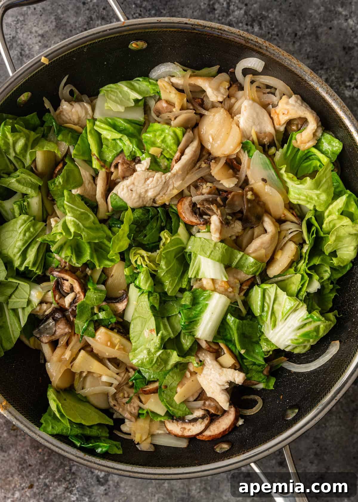 Process shot: Fresh bok choy leaves being introduced to the stir-fry in the same pan with chicken and other vegetables, showing the final stages of vegetable cooking for optimal freshness.