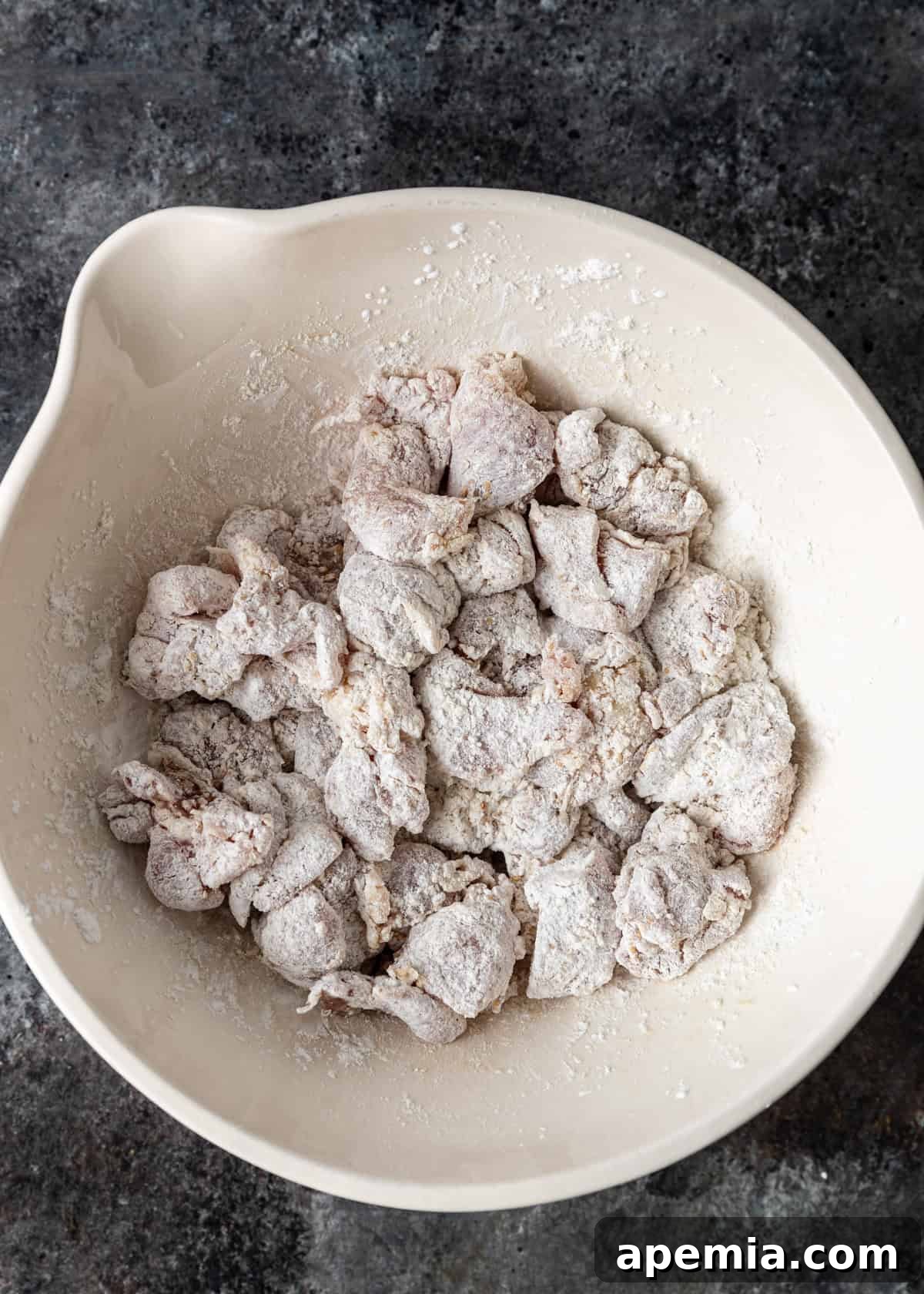 Overhead process shot: marinated chicken pieces being tossed with cornstarch and flour in a bowl, ensuring an even coating for General Tso's chicken.