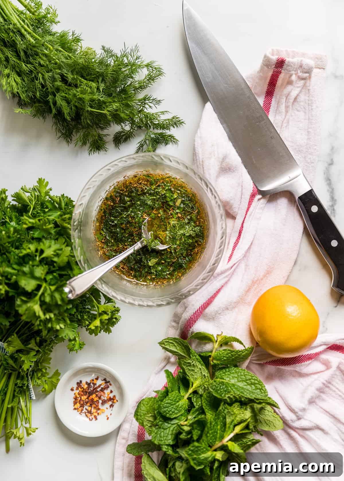Overhead view of fresh dill, parsley, basil, and a lemon alongside a dish full of Mediterranean chicken marinade ingredients.