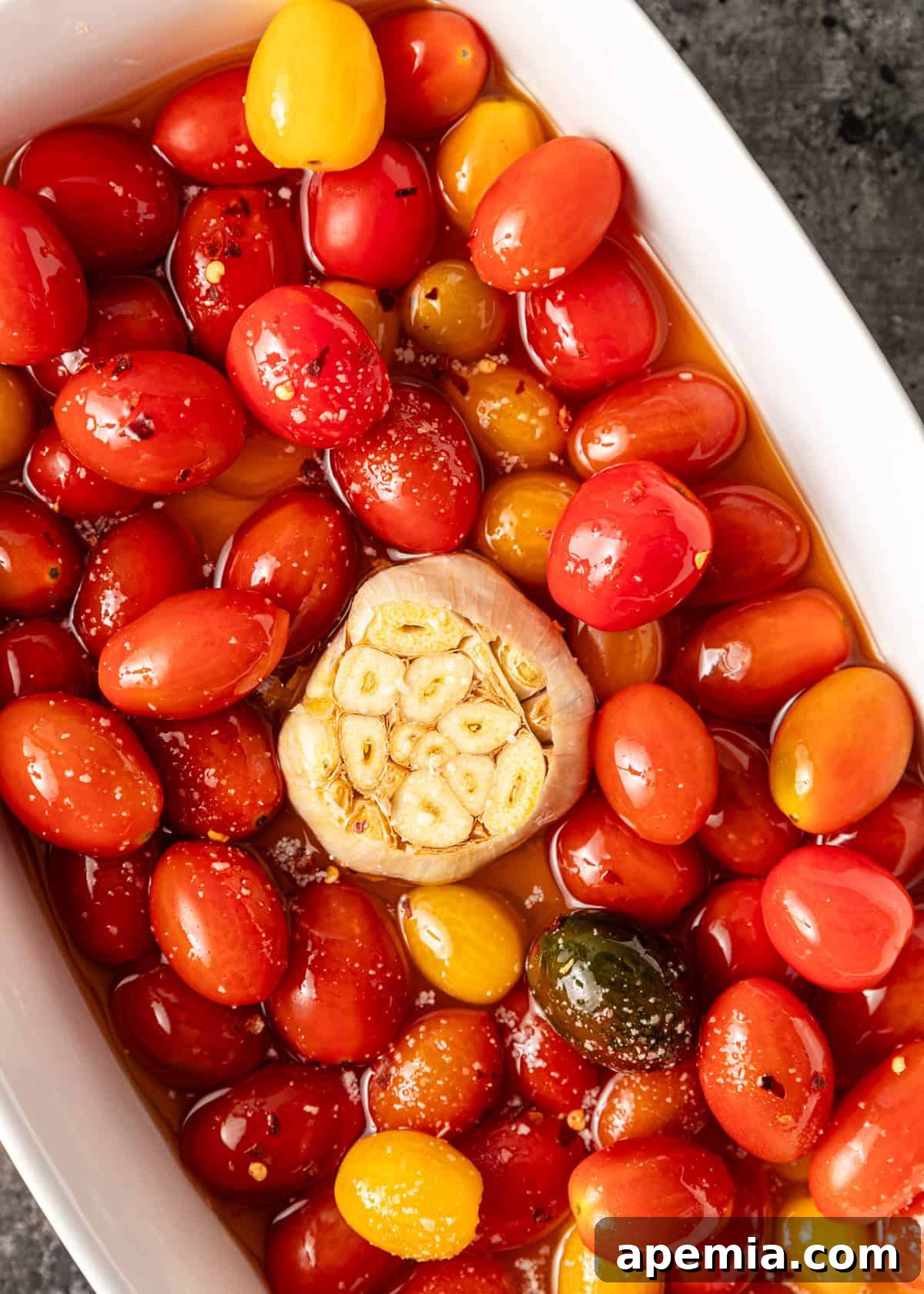 Overhead shot: cherry tomato confit in a baking dish before baking. Fresh tomatoes and garlic cloves submerged in olive oil.
