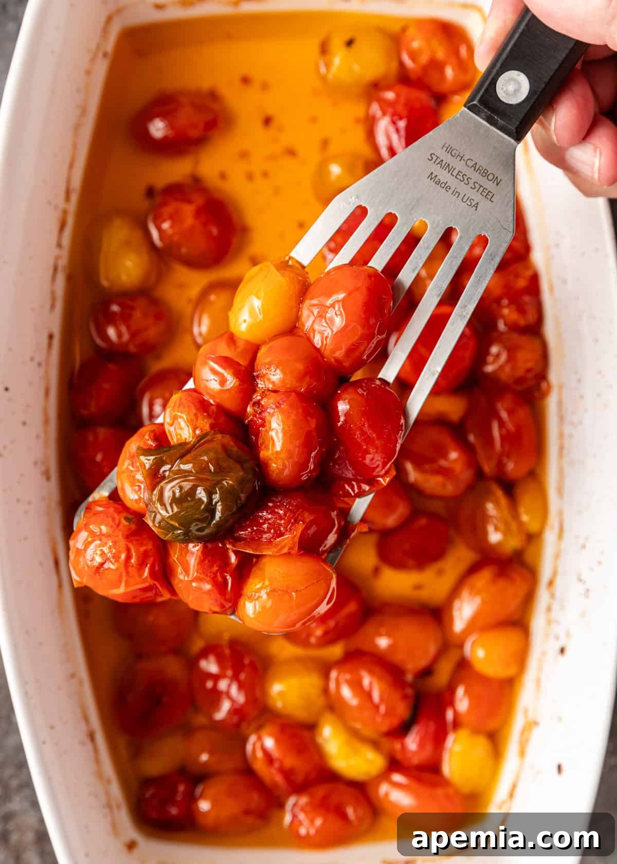 Overhead view of cherry tomatoes and garlic cloves undergoing the confit process in a baking dish, with a spatula lifting some